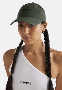 Cappellino da baseball in cotone verde scuro con logo RVCA nero, visiera curva, retro regolabile, indossato da una persona con treccine e una maglietta bianca.