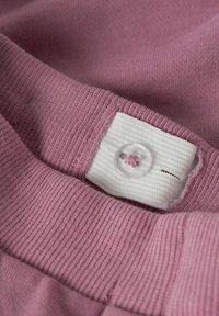 Lys rosa tekstil med en ribbet struktur, der har et hvidt stoffetiket og en gennemsigtig knap med en syet detalje.