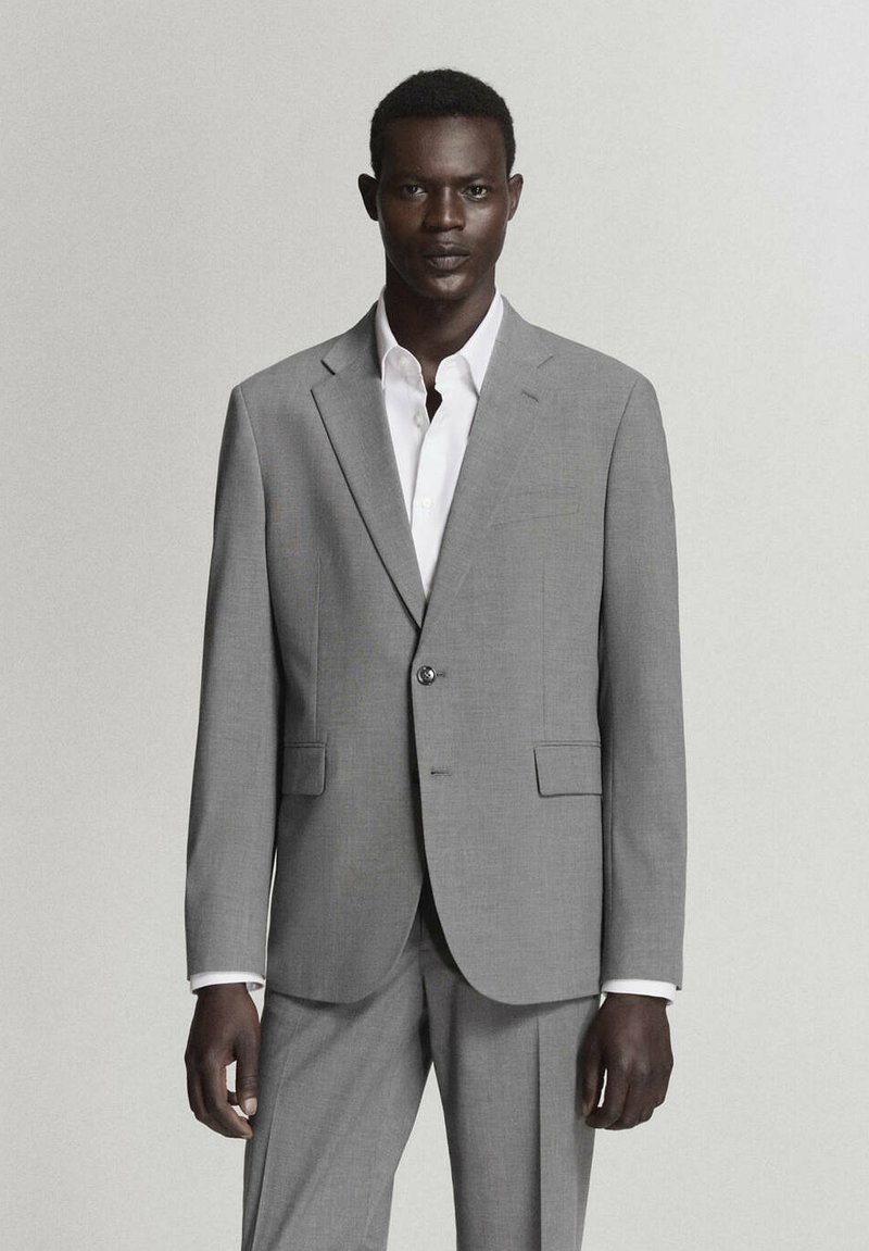 Homme noir debout, portant un costume gris clair avec une veste à un seul bouton et une chemise blanche sur un fond uni.