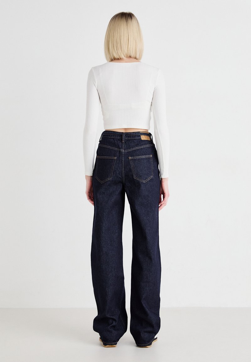 Jean en denim bleu foncé, à jambes larges et taille haute, avec des poches arrière visibles et une étiquette beige à la taille. Le modèle porte un haut court blanc.