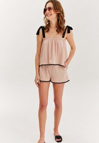 Žena oblečená v svetloružovom bezrukávovom overal s čiernymi okrajmi a ramennými viazaniami, slnečných okuliaroch, zlatých náhrdelníkoch a čiernych nazúvacích sandáloch.