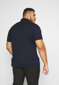 Navy polo shirt made of smooth fabric, featuring a pointed collar and short sleeves. Sleek fit with side slits at the hem, paired with black pants.