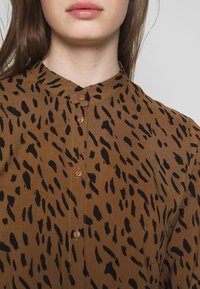 Braune Bluse mit einem schwarzen Tierdruckmuster, ausgestattet mit einer Knopfleiste vorne und einem Stehkragen. Glatte Textur, lockere Passform.