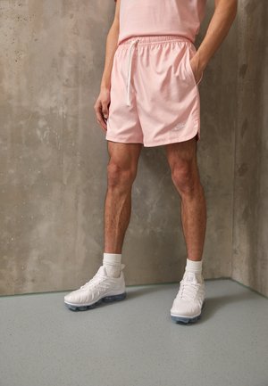 Osoba w jasnoróżowym T-shircie i szortach z białymi sneakersami stoi przy betonowej ścianie, ręce w kieszeniach, nogi widoczne.