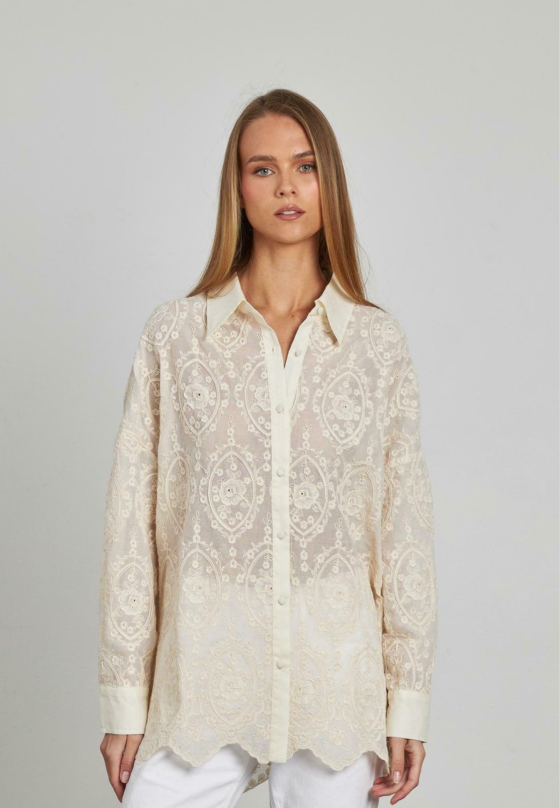 Durchsichtige Creme-Bluse mit aufwendiger floraler Stickerei. Knopfverschluss, spitzes Kragen und gewellter Saum. Lange Ärmel und weiche Textur.