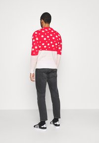 Hombre vistiendo un suéter de lunares rojos y blancos, jeans negros y zapatillas Adidas en blanco y negro de pie contra un fondo blanco liso.