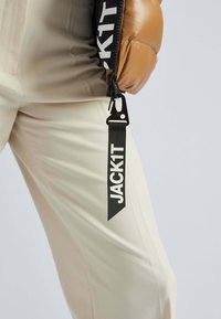 Giubbotto puffer beige con zip nera e grande testo "JACK1T" su una fascia. I pantaloni color crema completano l'outfit.