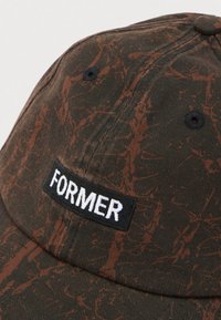 Σκούρο καφέ καπέλο του μπέιζμπολ με σχέδιο από κηλίδες σε σκουριά χρώμα και μαύρο μπατς με την επιγραφή "FORMER" σε λευκά γράμματα στην μπροστινή πλευρά.