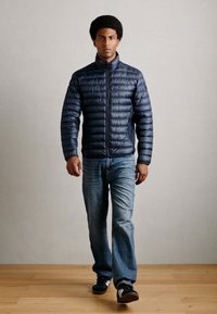 Marineblå pufferjakke med horisontal søm, glidelås og sidelommer, kombinert med blå jeans og svarte joggesko. Modell som går.