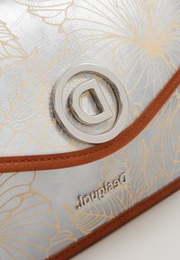 Srebrna syntetyczna torba z złotym kwiatowym wzorem, brązowymi skórzanymi akcentami i okrągłym srebrnym logo z metalowymi elementami. Gładka tekstura z błyszczącym wykończeniem.