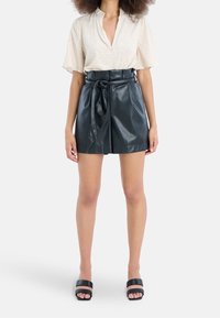 Černé šortky z umělé kůže s vysokým pasem a zavazovacím páskem, doplněné krémovou blůzou s krátkým rukávem a jemnou texturou. Černé nazouvací sandály.