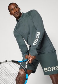 Σκοτεινό γκρι αθλητικό μπλούζα μακρυμάνικη με μισό φερμουάρ και λευκό λογότυπο "BORG" στους μανίκους, συνδυασμένο με αντίστοιχα σορτς και κρατώντας ένα ρακέτα τένις.