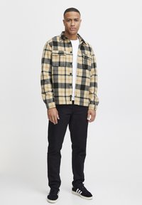 Checkered flannel shirt in yellow, black, and grey with a pocket; worn over a white T-shirt. Paired with dark trousers and black sneakers.