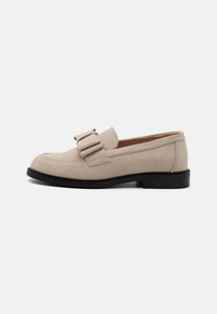 Beige nubuk bőr loafer, lekerekített orral, két díszcsattal, fekete gumitalppal és varrott részletezéssel a szélein.