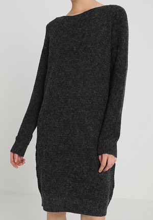 Vrouw draagt een donkergrijze, gebreide trui-jurk met lange mouwen tegen een effen lichte achtergrond.