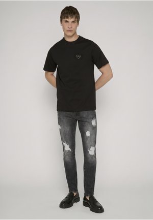 Jeune homme aux cheveux courts, debout les mains derrière le dos, portant un t-shirt noir, un jean gris déchiré et des mocassins noirs sur un fond uni.