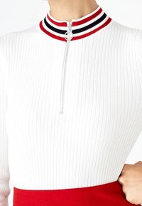 Haut blanc côtelé à col montant avec des rayures rouges et noires, fermeture éclair au niveau de l'encolure, associé à un bas rouge.