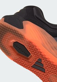 Sole til svart sportsko med teksturert mønster i oransje gummi. Har en svart sekskantet detalj og merkevarelogo på siden.