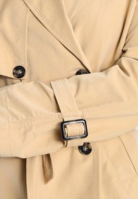 Manteau trench beige en gros plan sur la manche, mettant en avant des boutons noirs, une sangle ceinturée et une texture de tissu douce et lisse.