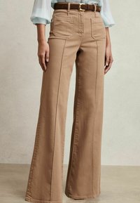 Pantaloni largi din material bej deschis, cu buzunare frontale, cusături contrastante și o curea maro cu cataramă aurie la talie.