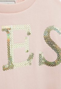 Primo piano di un tessuto rosa con lettere dorate in sequins luccicanti "E" e "S" su un indumento con scollatura a coste.