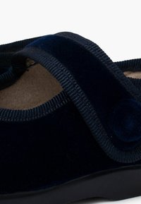 Zapato de terciopelo azul marino con una correa texturizada y un forro interior suave. Presenta una forma redondeada y una suela negra sólida.
