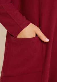 Hand met natuurlijke nagels gestoken in de zak van een lange mouw, zacht aanvoelend, donkerrode kleding.