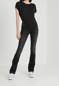 Black, flared jeans with a smooth texture, worn with a short-sleeved black top featuring white polka dots and white sneakers.
