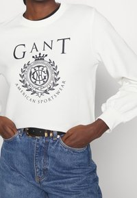 Felpa bianca con marchio GANT e logo grafico, abbinata a jeans denim blu e cintura nera con fibbia dorata.