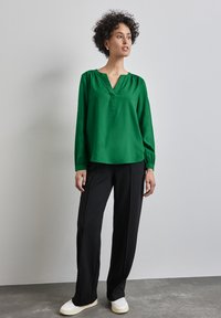 Blusa verde a maniche lunghe con scollatura a V e spalle arricciate; abbinata a pantaloni neri a gamba larga e sneakers bianche.