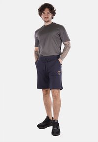 T-shirt grigio a maniche corte abbinato a pantaloni corti blu scuro con logo ricamato; indossato con scarpe da ginnastica nere.