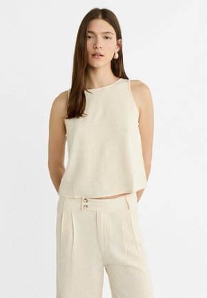 Top beige senza maniche con collo rotondo, realizzato in un tessuto texturizzato. L'orlo è leggermente curvato. Abbinato a pantaloni chiari a pieghe.