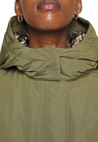 Chaqueta acolchada verde oliva con un cuello alto, textura suave y forro con patrón floral visible en el cuello.