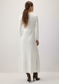 Witte geribde lange jurk met lange mouwen, aansluitende lijfje, uitlopende rok en een lengte tot halverwege het kalf. Gestructureerde stof; gedragen met bruine enkellaarsjes.