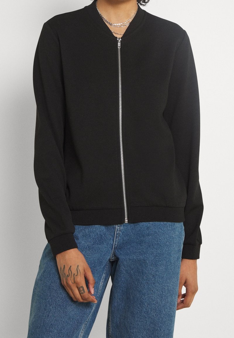 Personne portant une veste noire zippée, un jean bleu clair et des colliers argentés, avec un tatouage de flamme sur la main gauche.