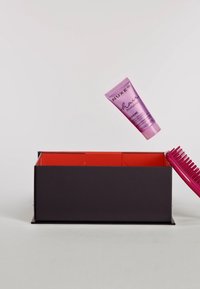 Lila hårkräm i tub märkt "NUXE hair" ovanför en svart låda med röd insida, bredvid en ljusrosa kam med rundade tänder.