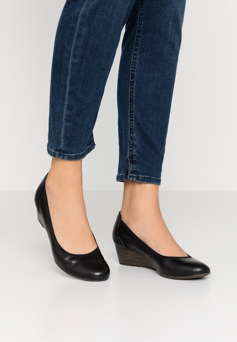 Pieds portant des compensées en cuir noir associées à un jean court bleu foncé sur un sol gris clair.