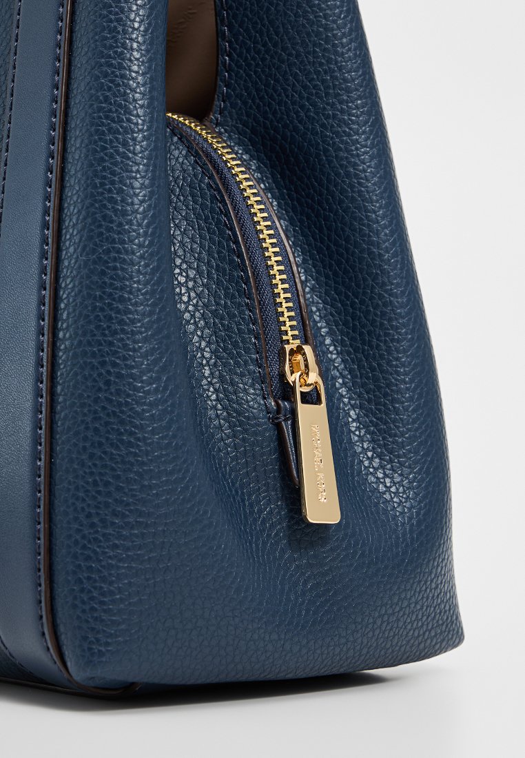 Sac à main en cuir texturé bleu marine avec quincaillerie dorée, comportant une fermeture éclair proéminente et une étiquette affichant le nom de la marque.