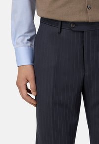 Jūras zila svītraina bikses, izgatavotas no gludas auduma, ar priekšējo pogu aizdari, virs tām redzama gaiši zila krekla piedurkne.