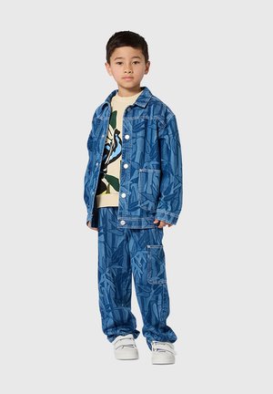 Blaue Jeansjacke und passende Hose mit Blattmuster. Das Outfit wird mit einem beigefarbenen T-Shirt mit Grafik und weißen Turnschuhen kombiniert.