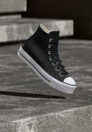 Schwarzer High-Top-Sneaker mit weißer Kappe und Sohle, schwebt über Betonstufen im urbanen Außenbereich.