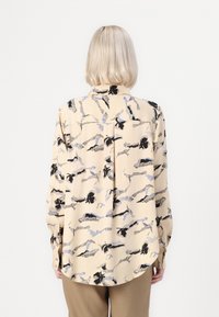 Long-sleeve blouse in light beige with black and blue abstract pattern. Features a collar, buttoned cuffs, and a relaxed fit.