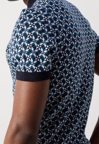 Zbliżenie osoby w koszulce z krótkim rękawem w niebiesko-biało-czarny geometryczny wzór oraz ciemnymi prążkowanymi mankietami.