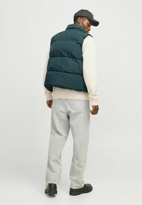 Donkergroen puffer gilet met een hoge kraag, dat drie horizontaal gestikte secties toont. Gedragen over een lichte trui en lichte spijkerbroek.