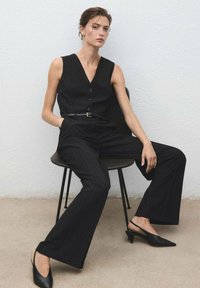 Black pinstripe vest and wide-leg trousers, paired with black slingback heels, seated on a black chair against a textured white wall.
