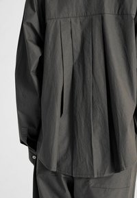 Camicia plissettata grigio scuro con vestibilità ampia. Presenta un tessuto liscio e texturizzato e bottoni sui polsini. Le fessure laterali migliorano il movimento.
