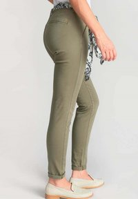 Olijfgroene, slim-fit broek met een gladde textuur, voorzakken en een opgerolde zoom. Vergezeld van een gestreepte sjaal die om de taille is geknoopt.