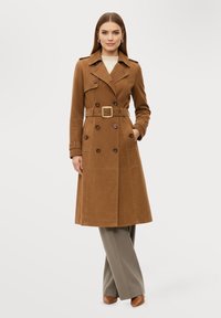 Kvinna klädd i en brun trenchcoat med skärp över ljusgrå byxor och bruna spetsiga skor, stående med ena handen i fickan.