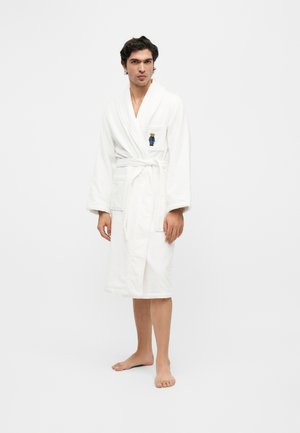 Homme debout pieds nus portant un peignoir blanc mi-long avec une ceinture nouée et une broderie d'ourson sur la poche poitrine.