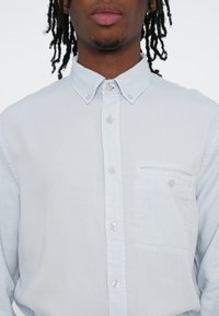 Světle modrá košile na knoflíky z hladké látky, s límečkem, jednou náprsní kapsou a rovně střiženým lemem.